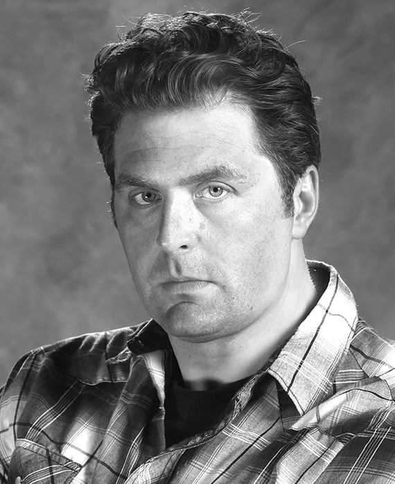 Black and white headshot of Elwood Reid in plaid flannel with gradient, marble background