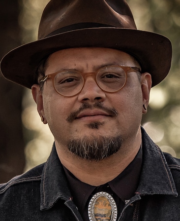Headshot of executive producer Sterlin Harjo of FX's The Lowdown, wearing bolo tie, round glasses and a brown bowler hat.