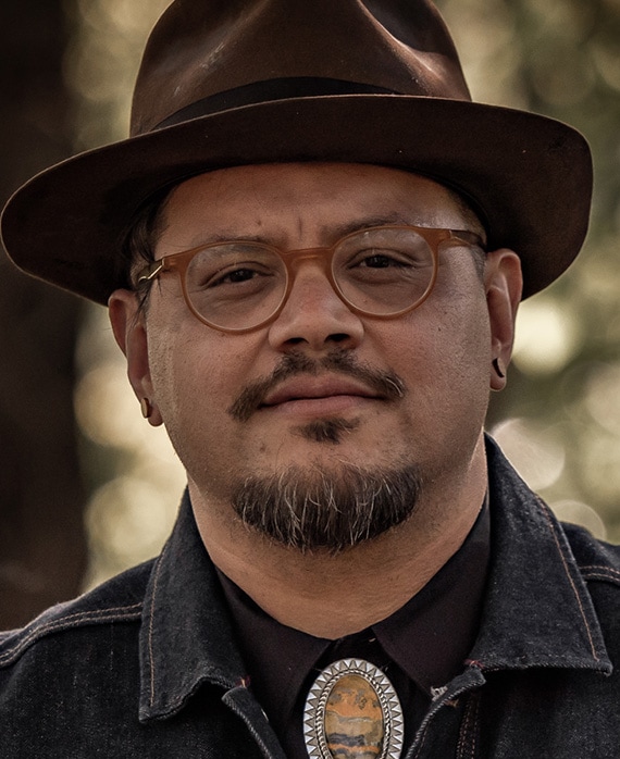 Headshot of executive producer Sterlin Harjo of FX's The Lowdown, wearing bolo tie, round glasses and a brown bowler hat.
