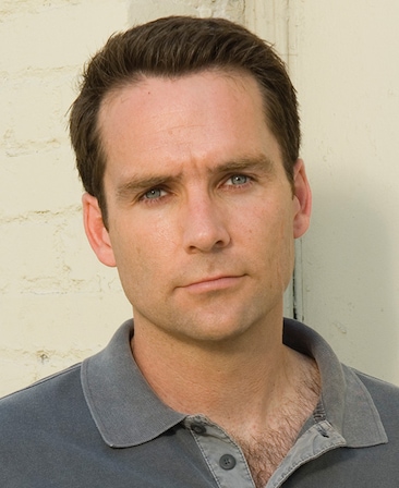 David Rees Snell headshot wearing a gray shirt with collar and buttons, leaning against white brick wall