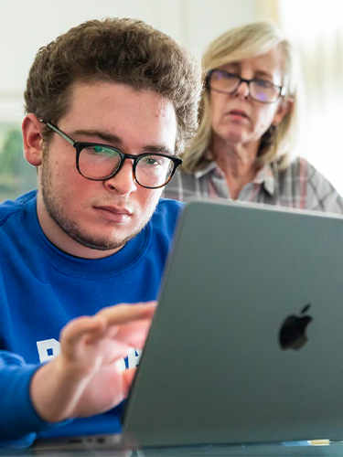 Boy with glasses and a blue sweatshirt typing on a laptop with a woman standing behind him.