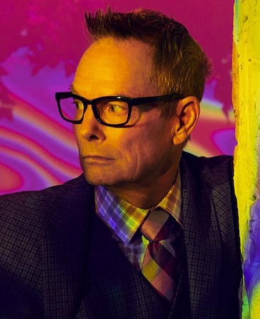 Bill Irwin headshot in black blazer with checkered multicolor tie, looking to the side with hallucinogenic purple background