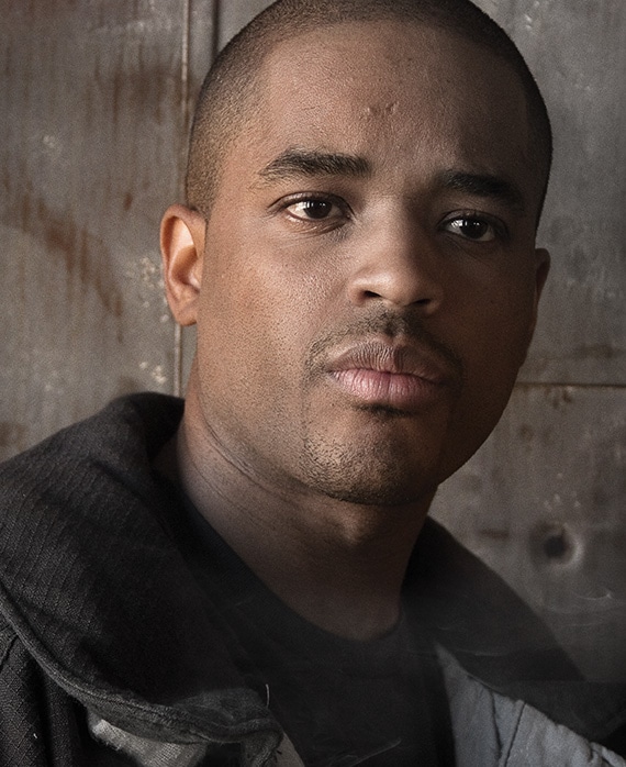 Larenz Tate headshot wearing a black jacket and standing in front of a metal wall