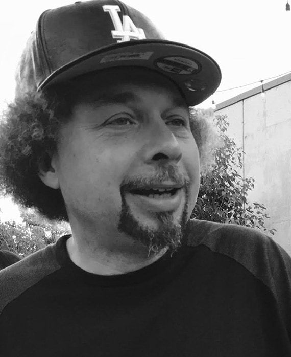 Headshot of executive producer Malcom Spellman wearing black and grey shirt and black hat.