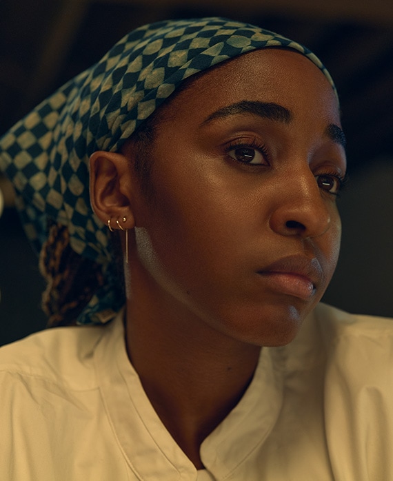 Ayo Edebiri headshot wearing a white chef jacket and a blue and white checkered bandana.