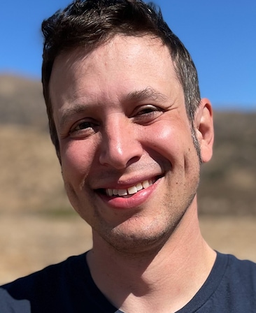 Headshot of executive producer Duffy Boudreau, wearing a blue t-shirt smiling outside near a small desert hill.