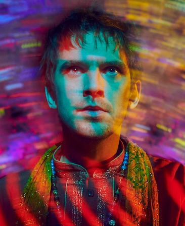 Dan Stevens headshot in red high neck top with yellow beaded scarf, with psychedelic multi color and blur effect