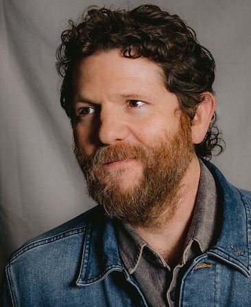 Headshot of Joshua Zetumer in a denim jacket with a grey background.