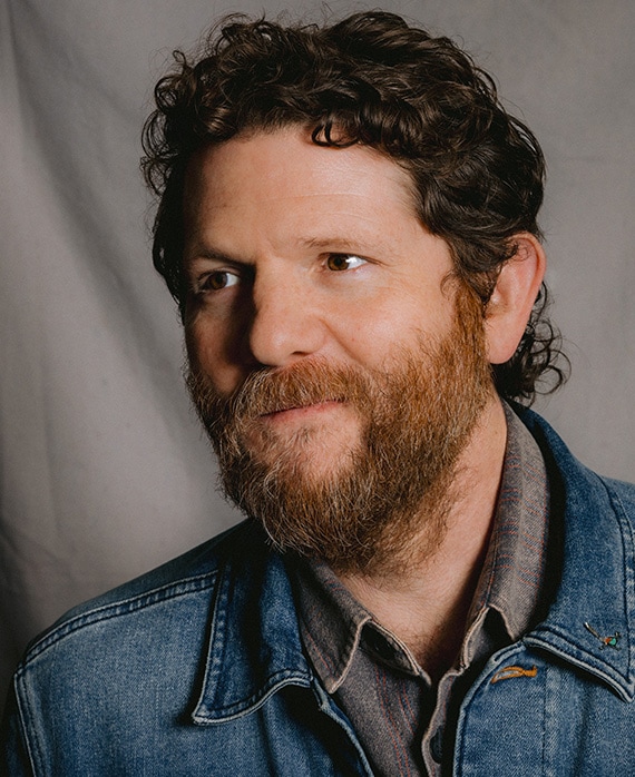 Headshot of Joshua Zetumer in a denim jacket with a grey background.