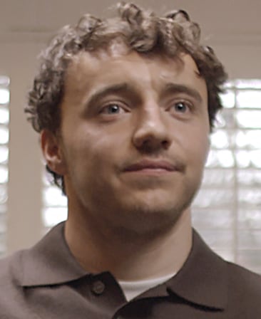 Ollie Rathbone headshot, wearing a brown button up with a white shirt peaking underneath, standing in front of window blinds.