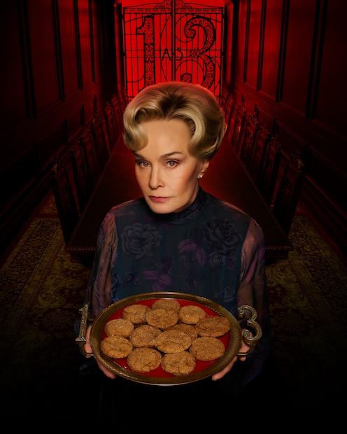 Jessica Lange is holding a platter of 13 cookies. The platter's handles have '13' on them, and she is in a dimly lit, dark red dining room with a gate that says '13' at the back.