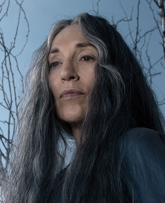 Katey Sagal headshot in dark colored top, looking down with wooden tall branches behind her with blue sky