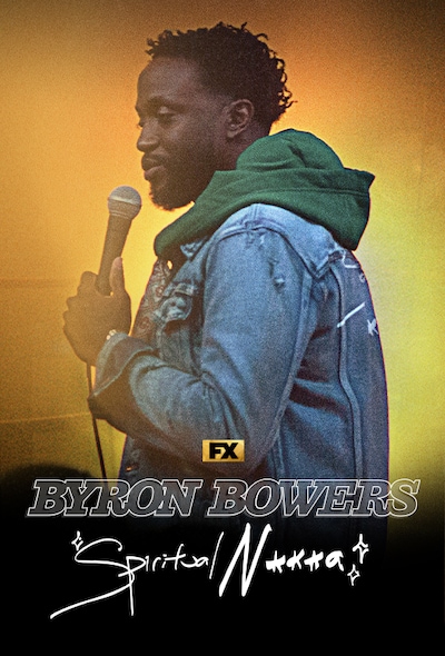Comedian Byron Bowers on a stage holding a mic wearing a denim jacket and green hoodie with a spotlight shining behind him