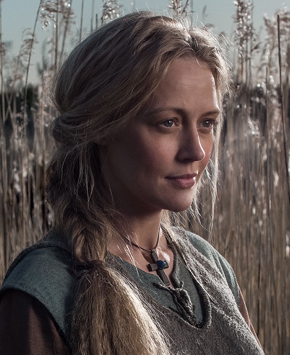 Elen Rhys headshot standing in wheat field with peasant blue and grey layered clothes with wooden cross necklace