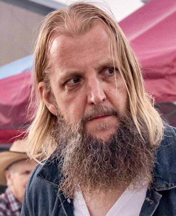 Headshot of executive producer Scott Teems wearing a white t-shirt, denim jacket outside near a red tent.