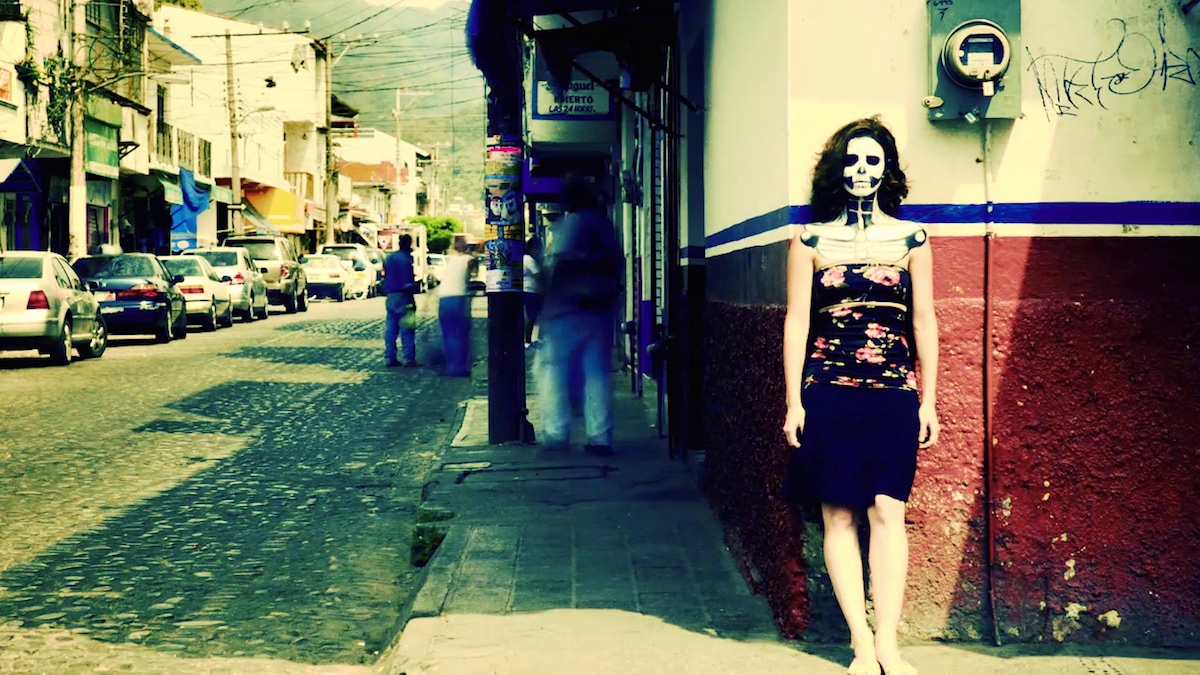 Woman in floral top and black skirt leaning against red wall by street with skull makeup on her face 