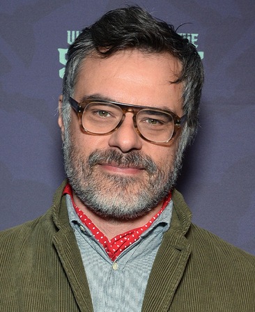 Jemaine Clement headshot wearing glasses with a green jacket.