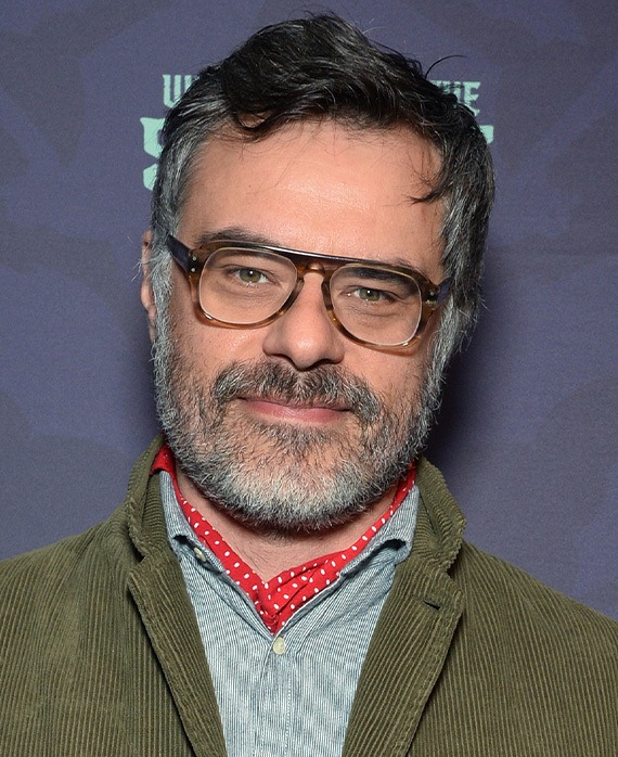 Jemaine Clement headshot wearing glasses with a green jacket.