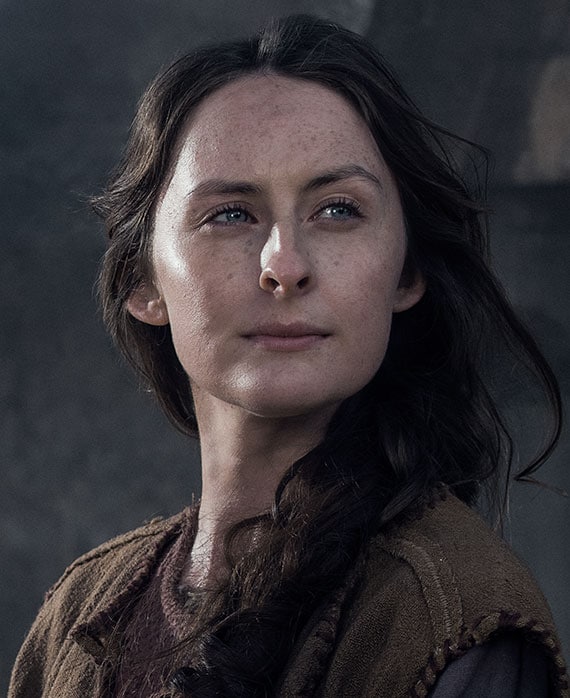 Sarah Sweeney headshot in layered brown ancient peasant clothing with hair braided in side ponytail