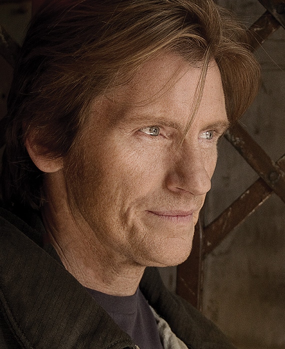 Denis Leary headshot wearing a black shirt and standing by a metal fence