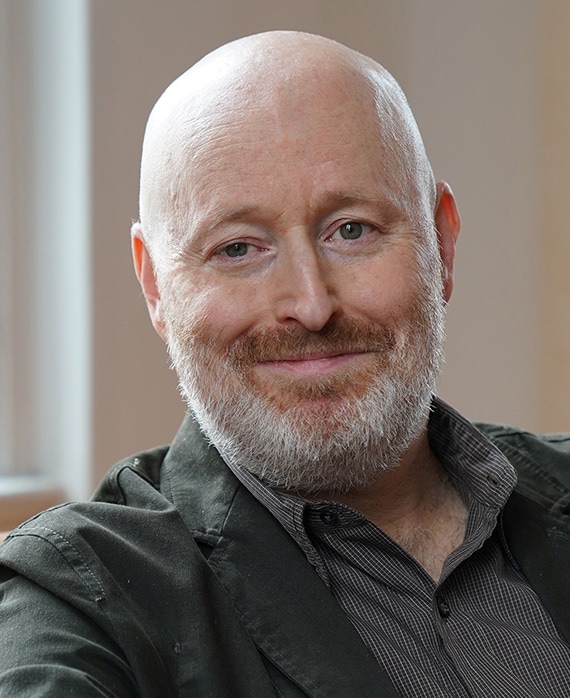 Joe Weisberg headshot in black and white patterned button down with thin black jacket with white wall and windowsill behind