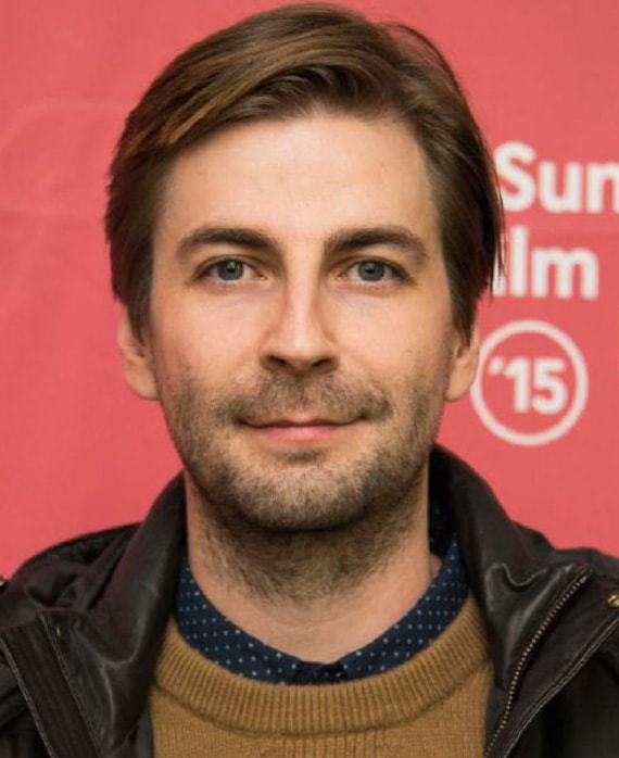 Jon Watts headshot wearing a brown sweater and black leather jacket standing in front of a red sign