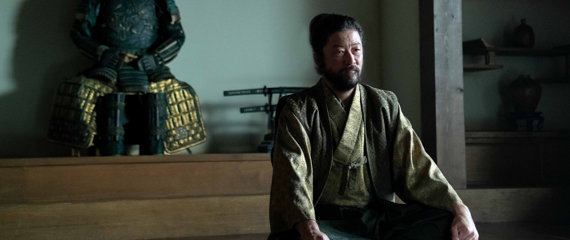 Yabushige dressed in a green kosode sits in his Edo meeting room in front of his armor