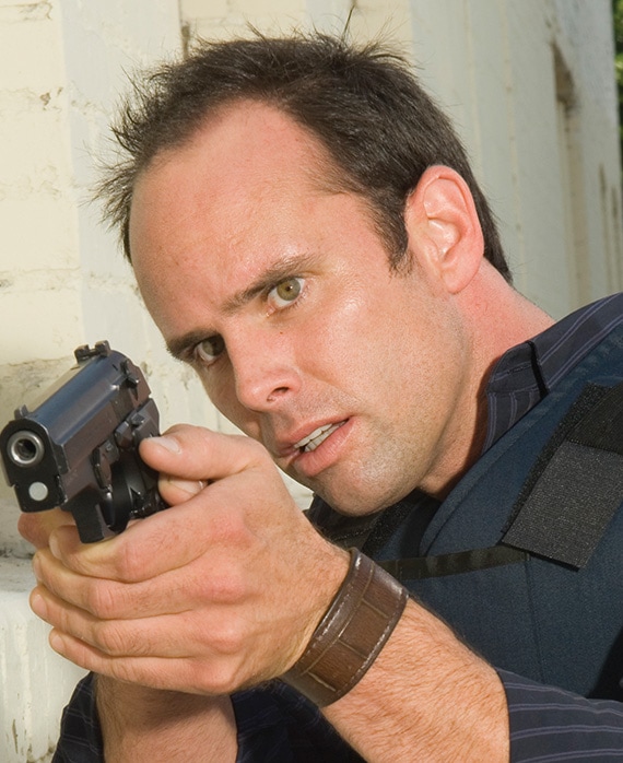 Walter Goggins headshot wearing a brown leather bracelet and body armor while holding a black gun