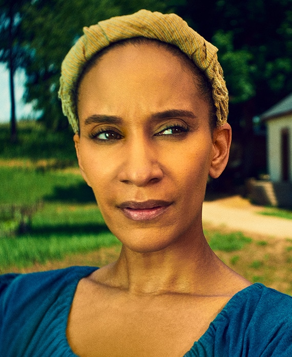Sophina Brown headshot in blue peasant top with yellow headband, with grass and trees in background with side of white shed