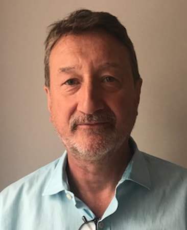 Steven Knight headshot in light blue button down with glasses hanging on shirt's button, with plain light brown background
