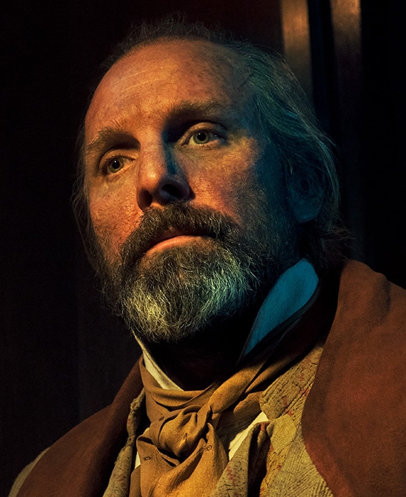 Johnny Harris headshot in brown suede vest with light brown neck tie scarf, looking up in dark room toward bright light