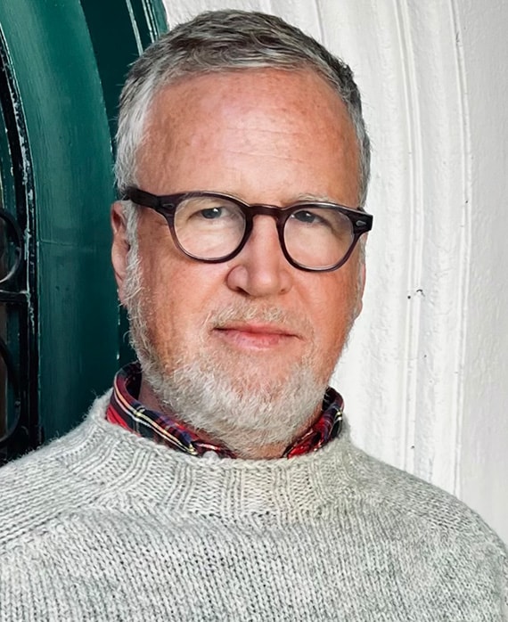 Ned Martel headshot wearing a grey sweater and brown glasses