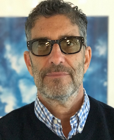 David Schiff headshot wearing black glasses with a blue plaid collar shirt and navy sweater