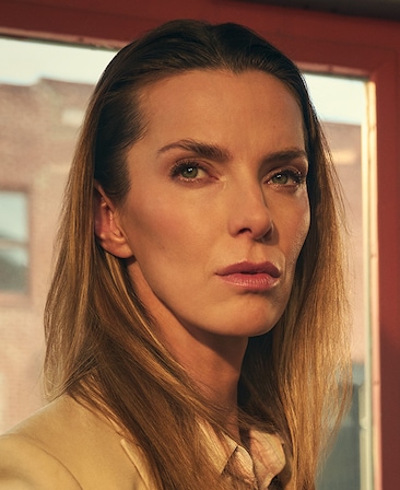 Headshot of Betty Gilpin as Ginger in FX's The Lowdown, wearing a tan suit and plaid button up, standing in front of a window.